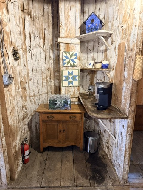 Small rustic corner with wooden walls and floor, featuring a wooden cabinet, shelves with decorative tiles, a coffee maker, a water bottle, a fire extinguisher, and a trash can.