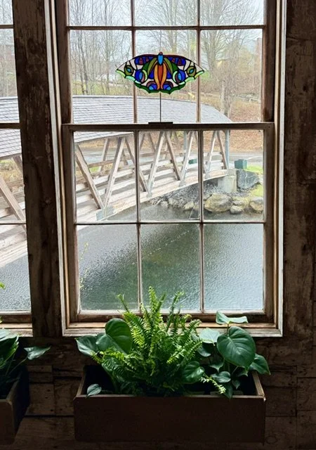 View of a pond and bridge through a window with stained glass butterfly hanging at the top.
