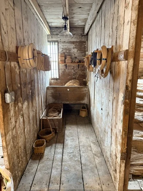 Wooden washroom with wooden barrels on the walls, a small window, and a basic light fixture. Contains baskets and a washbasin.