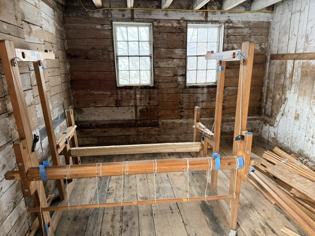 A woodworking loom frame inside an unfinished room with wooden walls, two windows, and a stack of wooden planks in the corner.