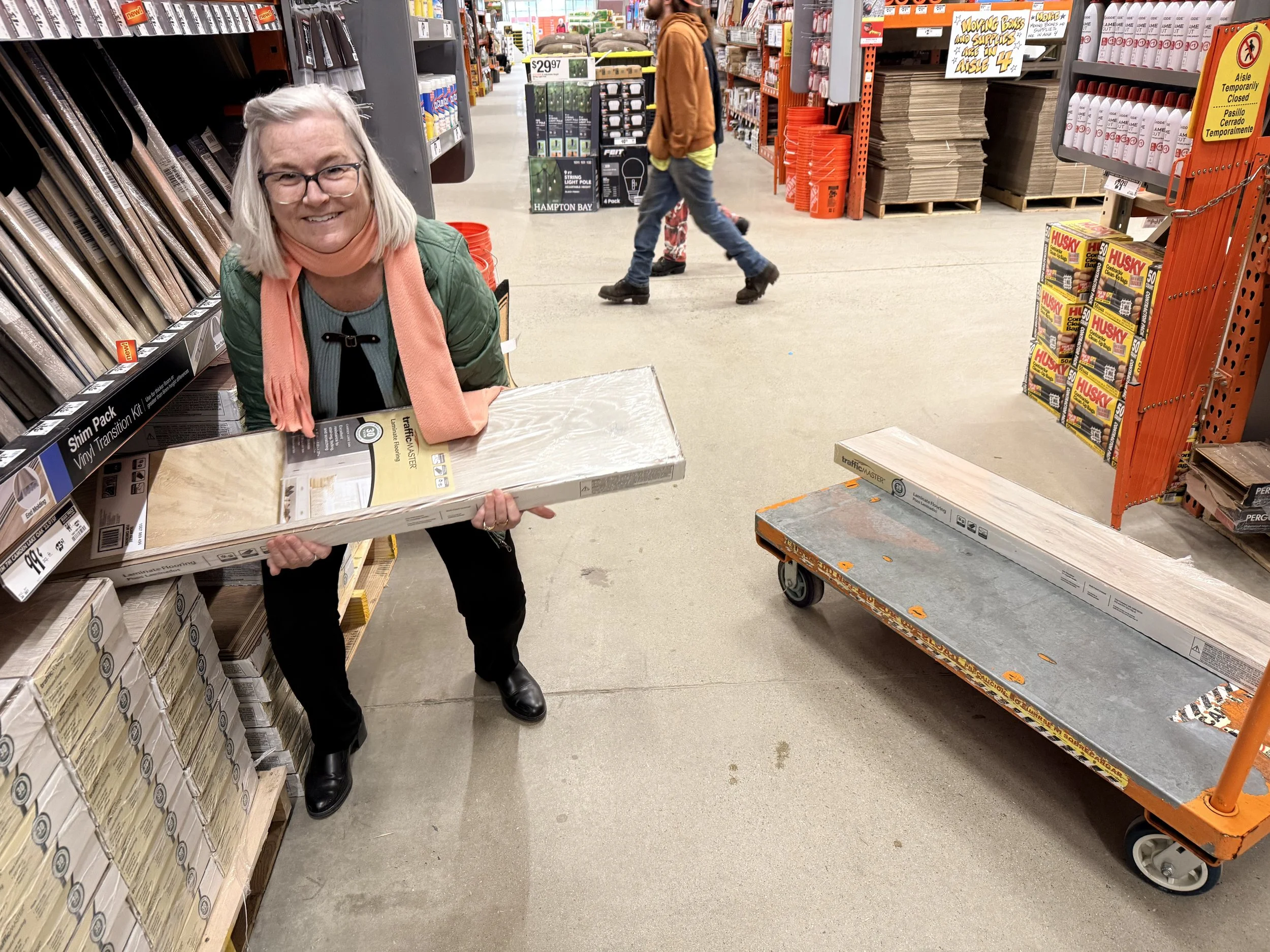 Sharon holding a boxed item in a home improvement store aisle, with shelves of building materials and other shoppers in the background.