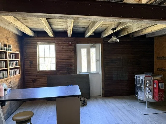 Interior of a rustic wooden kitchen or dining space with a table, a stool, a shelf with jars, a door, and a candy cart.