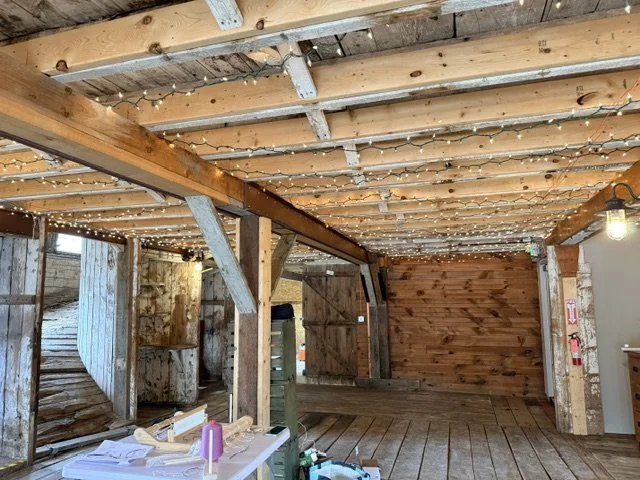 Interior of a rustic wooden barn or shed with exposed beams and string lights, wood walls, and a wooden floor.