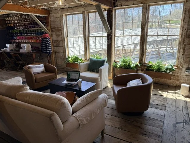Cozy rustic living room with beige and brown armchairs and a white sofa, a black coffee table with a laptop, large windows with a view of an outdoor wooden deck, and indoor plants on window sills.