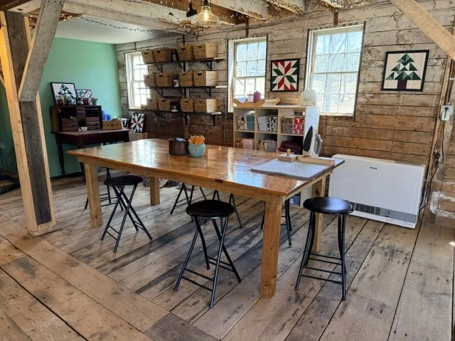 A rustic dining or craft room with a large wooden table and four black stools. The background features wooden walls, two large windows, and shelves with baskets and decorative items. There are quilts hanging on the walls and a small white bookshelf w