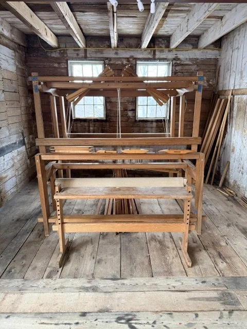 Stack of unfinished wooden furniture, including tables and racks, inside a rustic wood shed with two small windows.