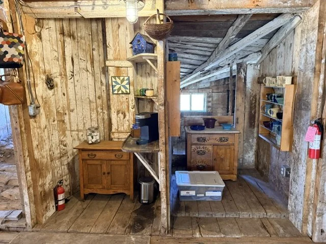 Interior of a rustic wooden cabin with various shelves and furniture, a window, a fire extinguisher, and kitchenware including bowls and containers.