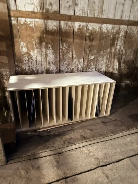 Wooden rack for sheets of stained glass with multiple compartments, placed against a weathered wooden wall in a rustic setting.