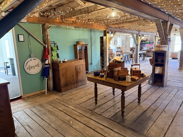 Interior of a rustic store or cafe with a wooden counter, a table with various items, and string lights on the ceiling