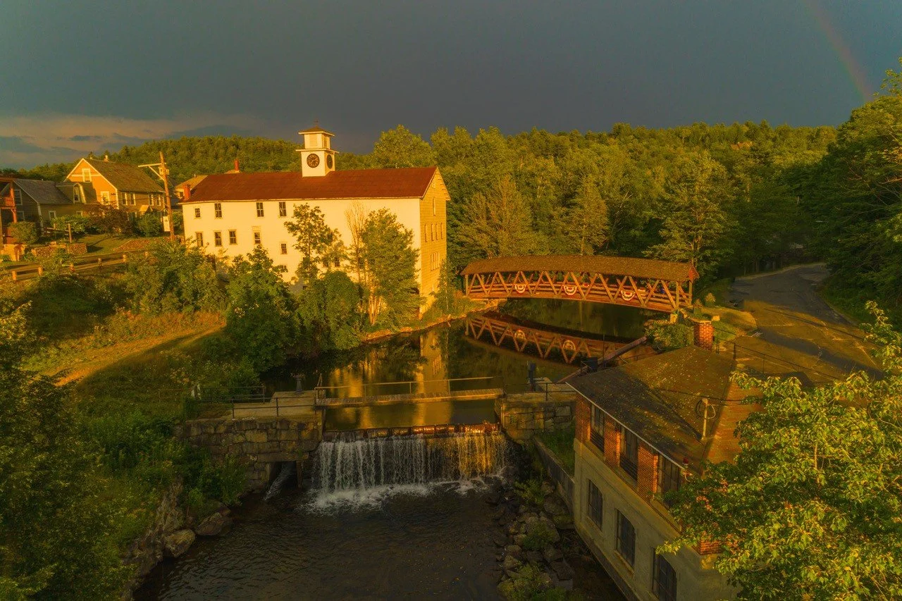A scenic view of a river flowing under a bridge and waterfall, with a historic white building with a clock tower, surrounded by trees and houses, under a dark sky with a rainbow.