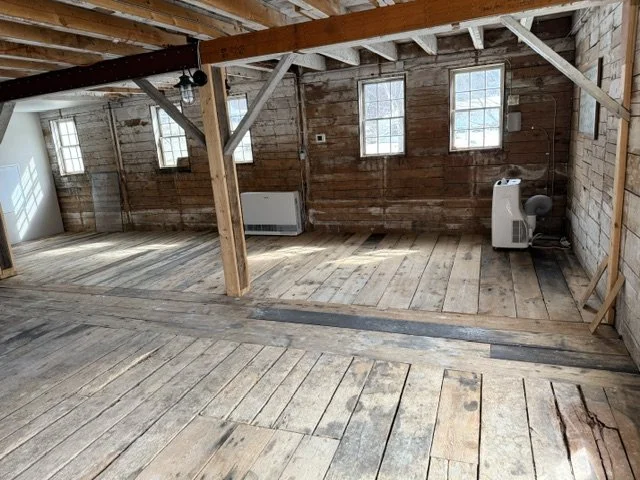 Interior of an unfinished wooden building with exposed beams, three windows, a radiator, and a portable heater.