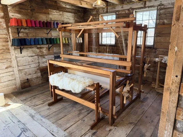 A wooden weaving loom in a rustic wooden room with multicolored yarn on shelves.