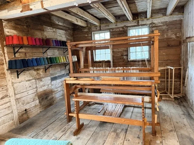 A wooden weaving loom set up inside a rustic wood-walled room with two small windows.