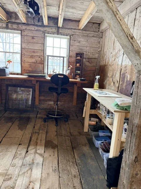 A rustic woodworking or craft space inside a wooden shed with two windows, a work table, a black rolling chair, and a small shelf with various supplies.