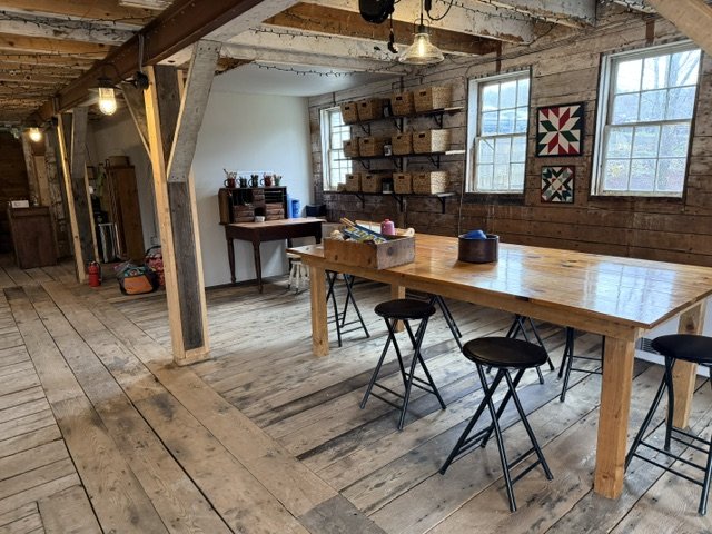 Interior of a rustic room with wooden walls and floors, featuring a large wooden table with four black chairs, open shelving with wicker baskets, and three windows letting in natural light.