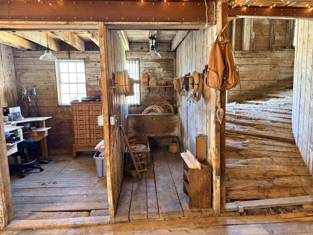 A rustic wooden workshop with tools and equipment, including a chair, shelves, and a backpack hanging on a wooden wall. The space has a lofted area with wooden stairs.