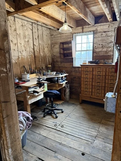 A rustic wood workshop with a workbench, a stool, a chest of drawers, and a window with light coming in.