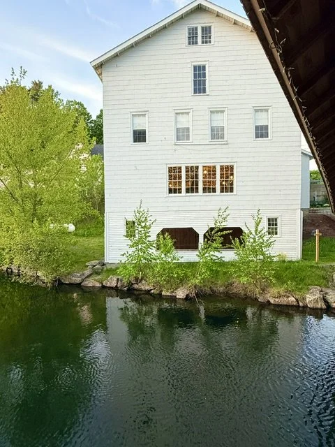 The back of the historic Sunapee Livery building, a white multi-story house next to a body of water, with trees and rocks along the shoreline.
