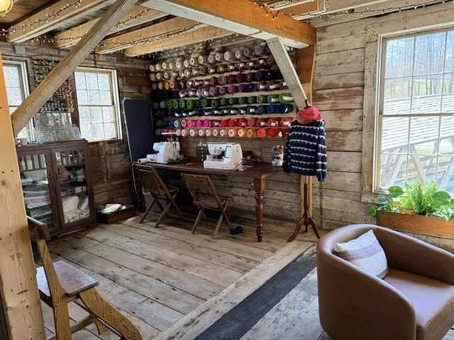 A cozy sewing room with wooden walls and floors, featuring colorful spools of thread on wall-mounted shelves, sewing machines on a wooden table, and a small plaid jacket hanging on a coat rack. There is a window with a window box of green plants, and