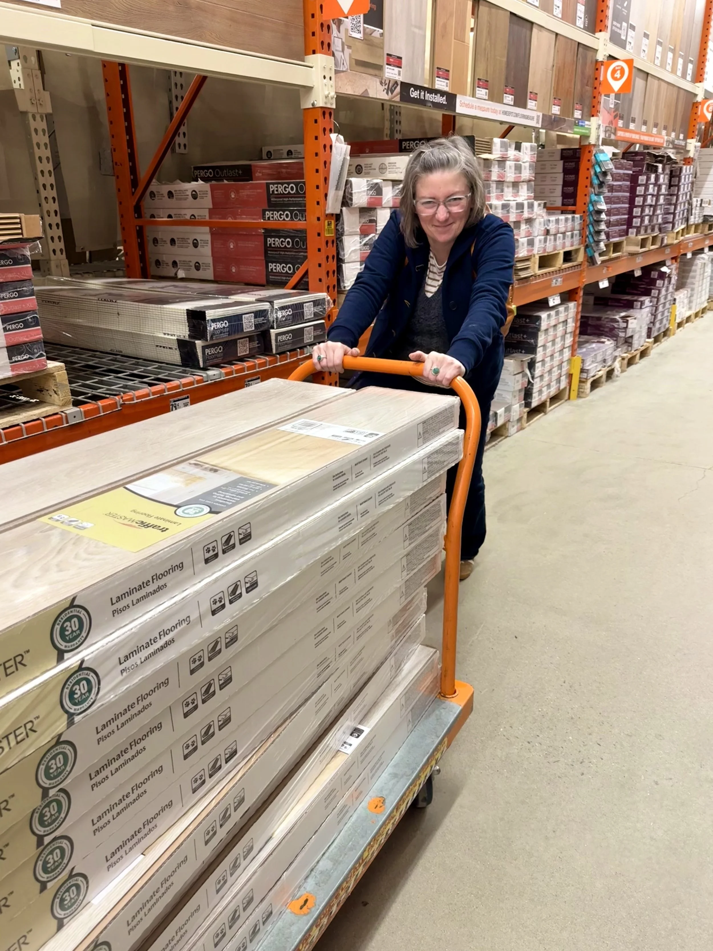 Katie shopping for flooring at a home improvement store, pushing a cart with boxes of laminate flooring visible in the foreground.