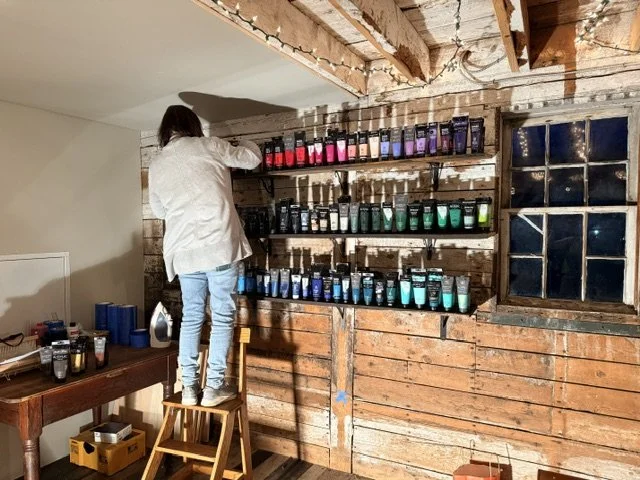 Person standing on a small wooden step stool arranging rows of colorful paint tubes on rustic wooden shelves in a room with exposed wooden beams and a window.