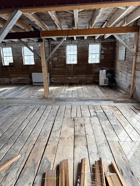 Interior of an unfinished wooden building with exposed beams, multiple windows, and a clear floor. Construction materials are on the floor in the foreground.