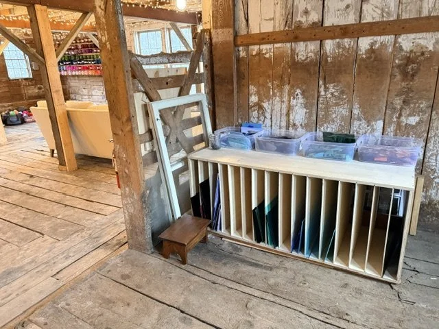 A wooden storage unit with vertical slots filled with stained glass, and transparent plastic containers on top, leaning against a rustic wooden wall in a barn-like space.