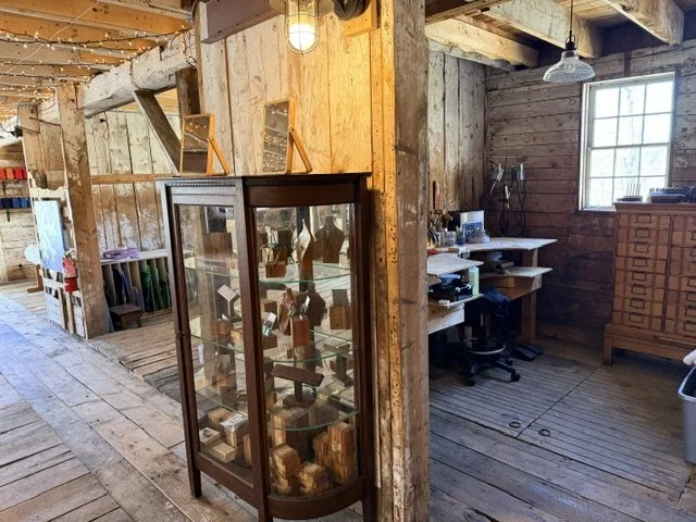 Display cabinet with small wooden figures inside, located in a rustic wooden interior with a work desk and window.