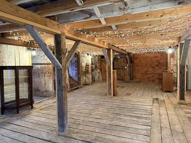 Empty rustic attic with wooden beams, string lights, and a wooden floor, featuring a glass display case and a small wooden cabinet.
