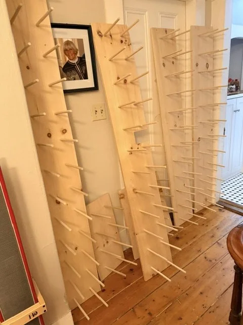 Three wooden display racks with multiple pegs in a hallway next to a white door and kitchen area.