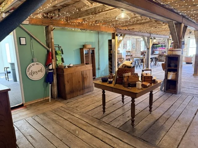 Interior of a rustic shop with wooden floors and exposed beams, decorated with string lights, featuring a checkout counter, a wooden table with various items, and seating area in the background.