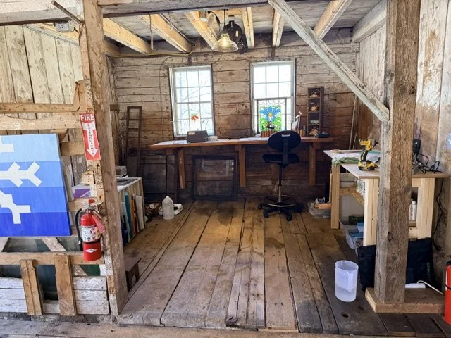 Interior of a rustic wooden cabin with a work desk by two windows, a black office chair, a small bookshelf, and various tools and supplies.