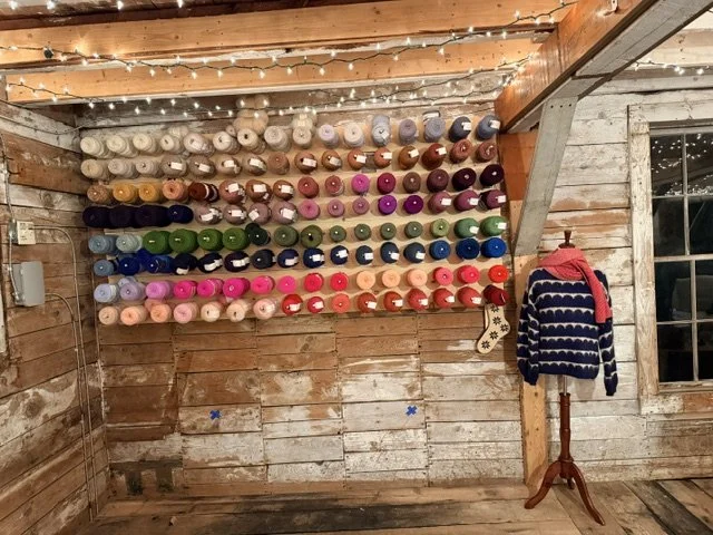 Wall of colorful yarn spools organized by colors in a rustic wooden room decorated with string lights, with a cozy sweater hanging on a wooden stand nearby.