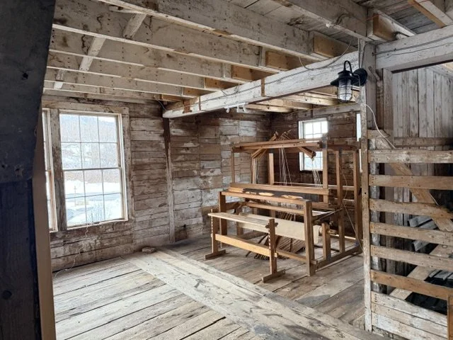 Interior of a rustic wooden building with exposed beams, wooden walls, and a wooden floor. There are two windows, one on the left and one on the back wall, with natural light coming through. A wooden loom and a mirror are placed against the back wall