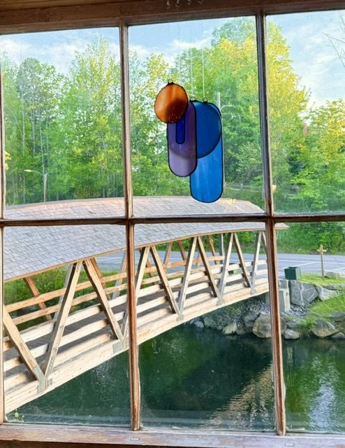Colorful glass art piece hanging in front of a window with a view of a wooden bridge over a pond and green trees outside.