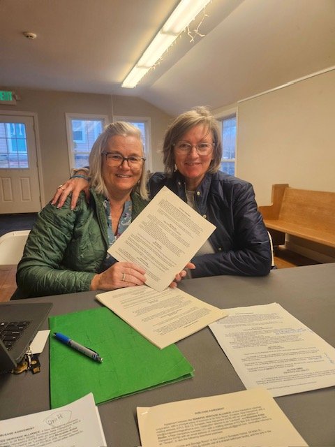 Sharon and Katie sitting at a table, smiling, holding documents, with papers and a marker on the table, in a room with windows and a door in the background.