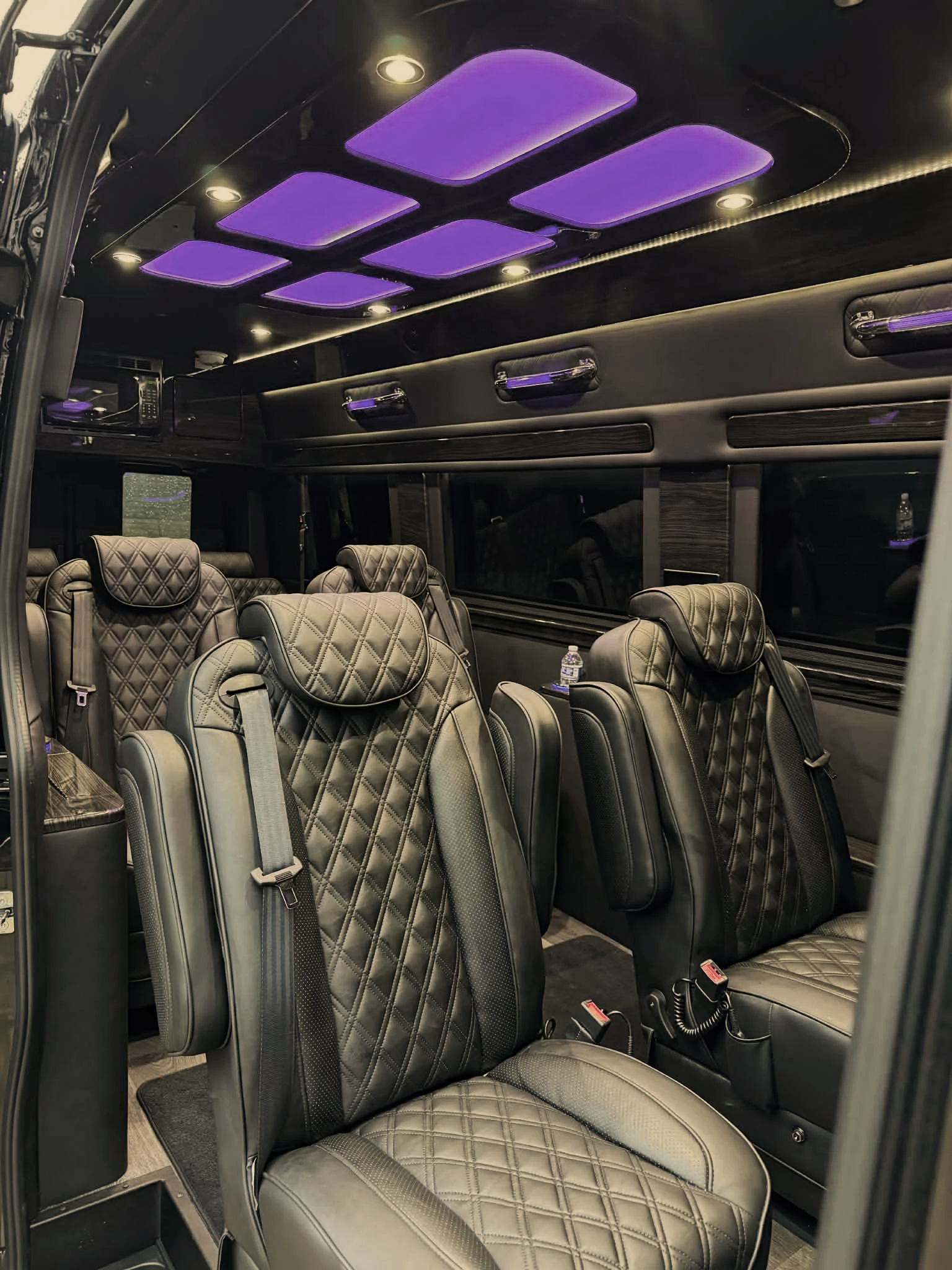 Interior of a luxury vehicle or limousine with black leather seats, overhead purple lighting panels, and small windows along the side.