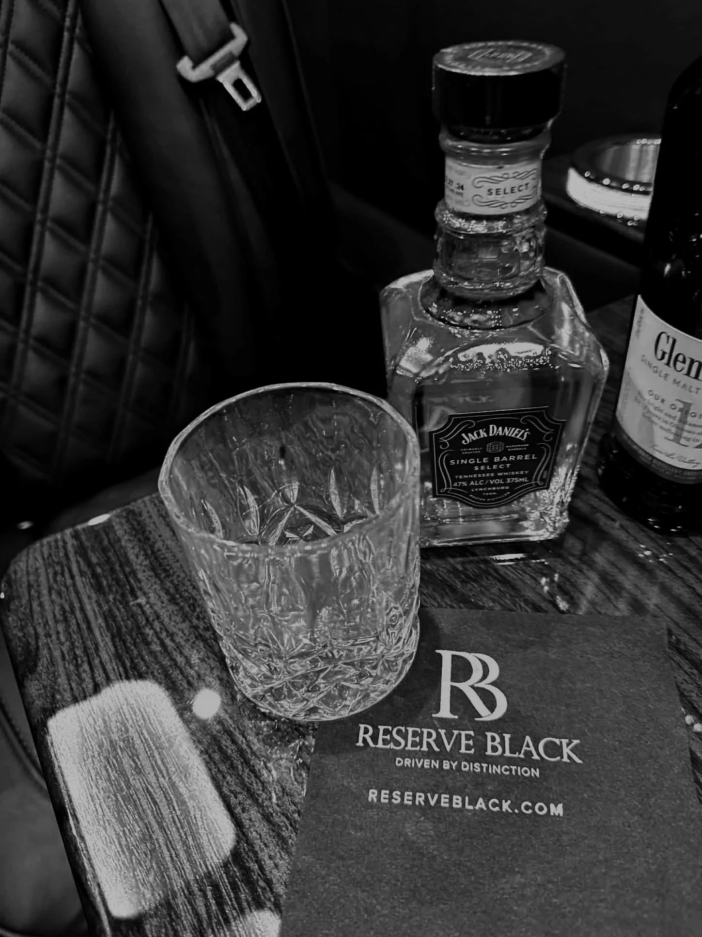 A glass of whiskey on a wooden table next to a bottle of Jack Daniels whiskey, a bottle of Glenfiddich whiskey, and a reservation black menu or card.
