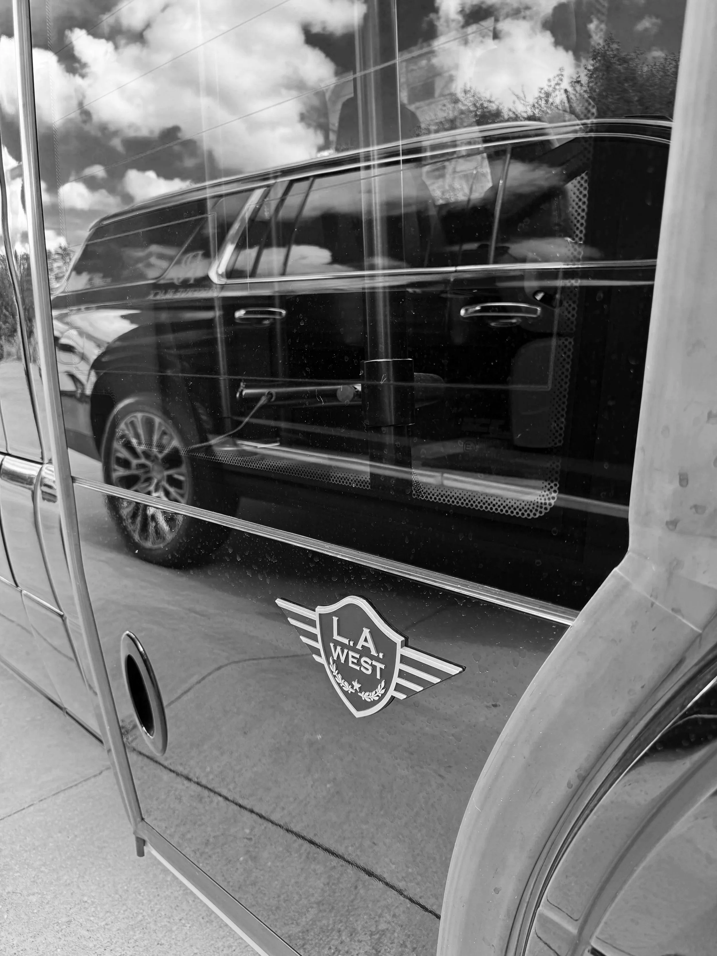 Black and white photo of a vending machine displaying a sticker with the logo 'L.A. WEST' on the lower part of the machine. Reflections of a car, sky with clouds, and trees are visible in the glass of the vending machine.