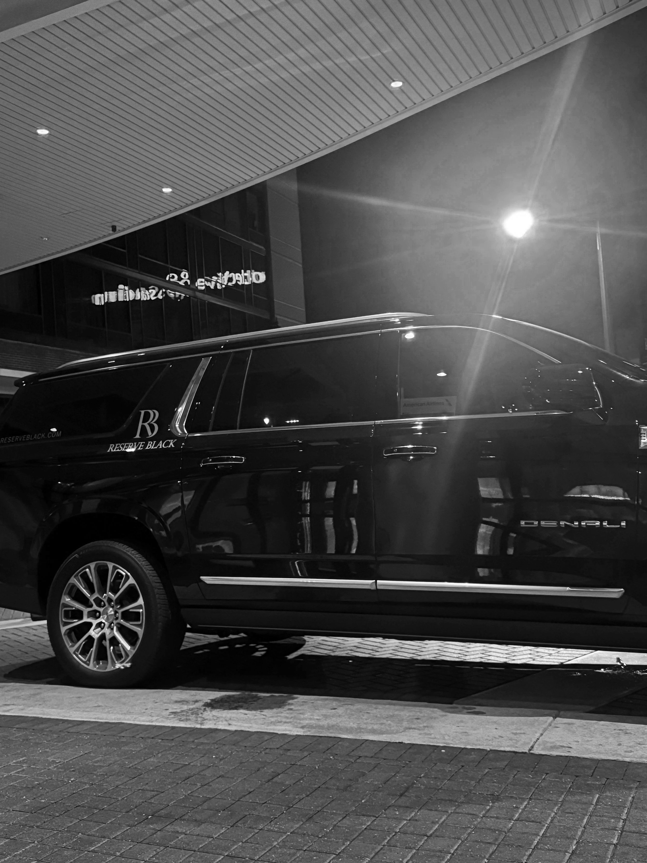 Black luxury car parked outside a modern building at night, with bright street lights and glass windows reflecting light.