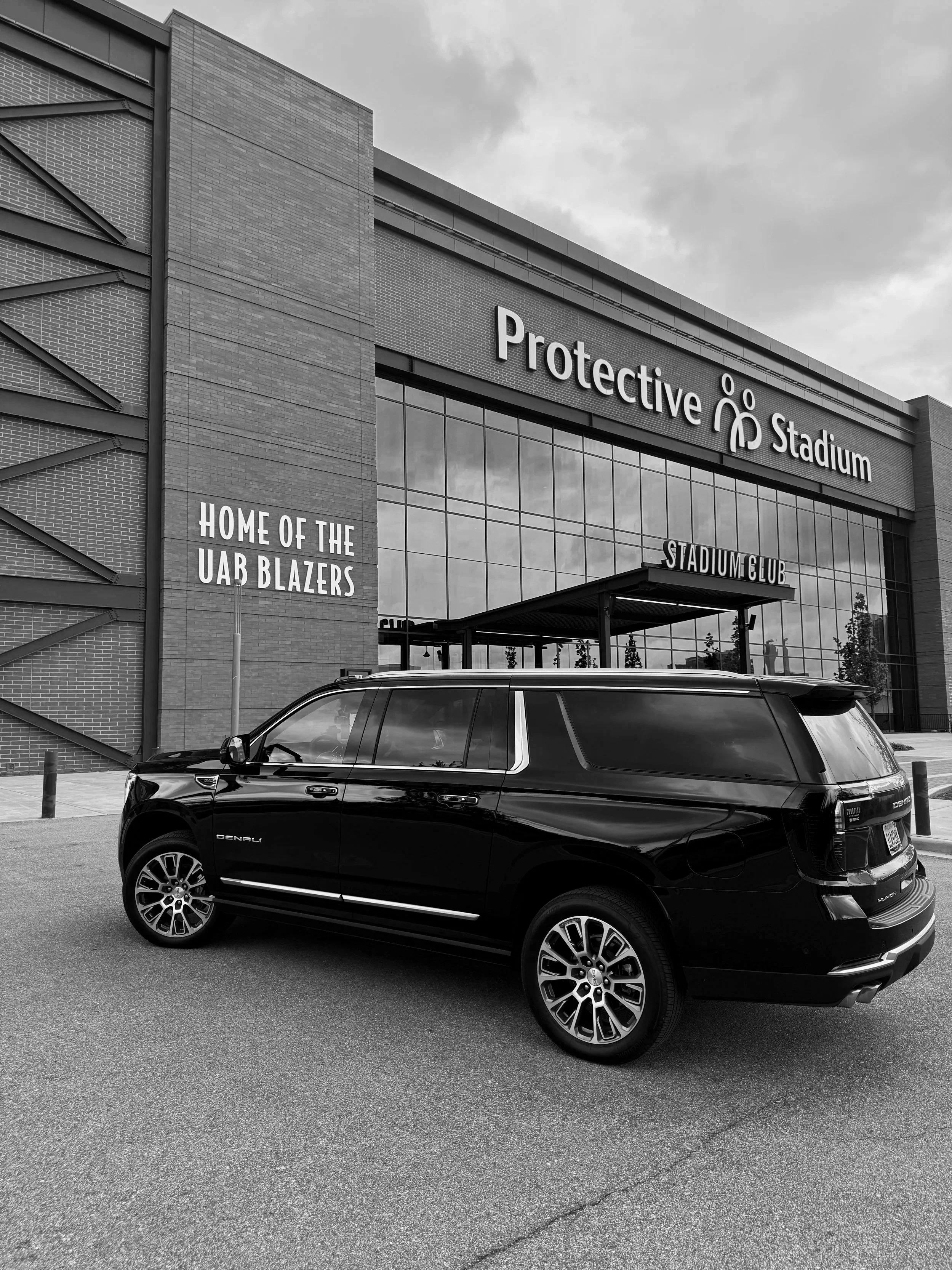 A black SUV parked in front of a large building with glass windows. The building has the signs 'Protective Stadium' and 'Home of the UAB Blazers'. The sky is cloudy.