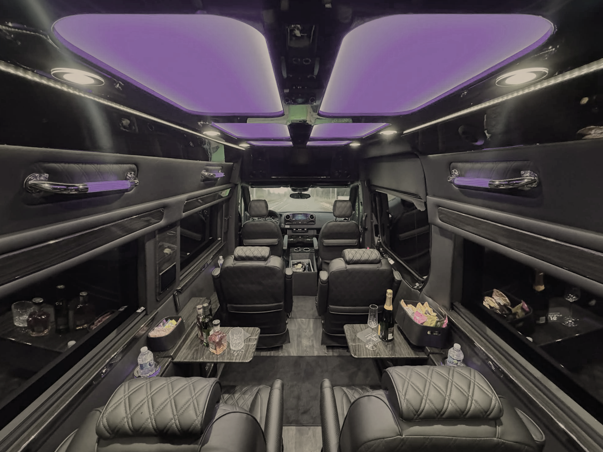 Interior of a luxury limousine with black leather seats, small tables with bottles and glasses, mood lighting with purple hues, and a view of the driver's cabin.