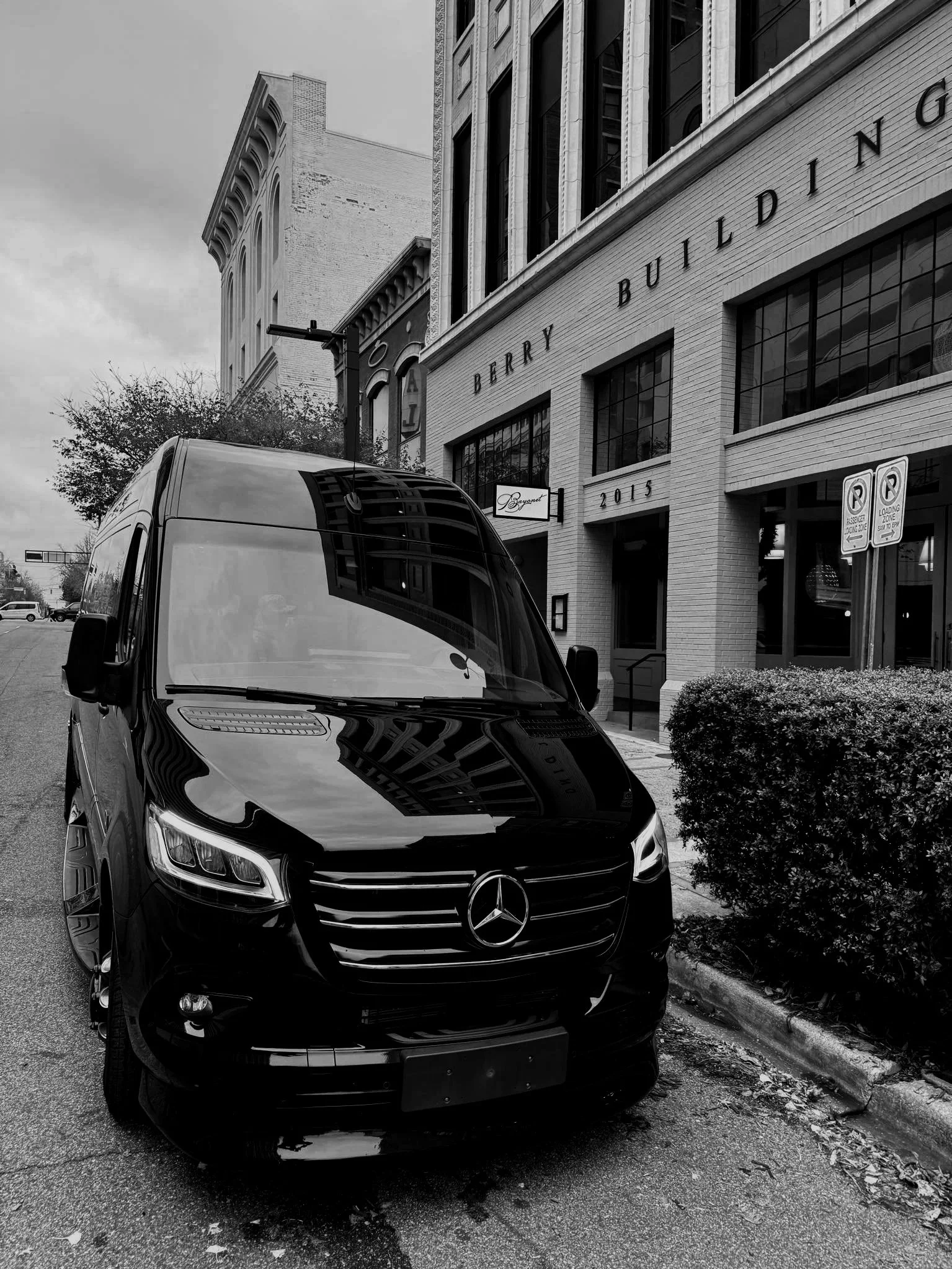 Black Mercedes-Benz van parked on city street near a building labeled 'Berry Building' with large windows and a sign for 'Bouquet.' The scene is in grayscale.