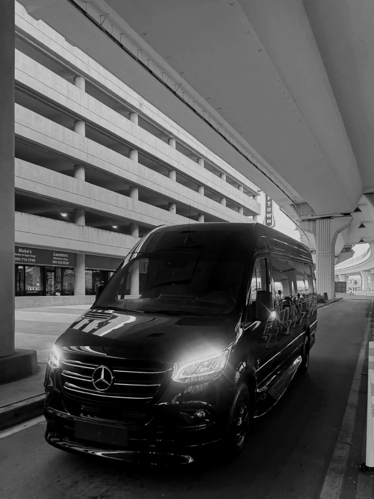 A black Mercedes-Benz van parked under an overpass near a multi-story parking garage with retail stores.
