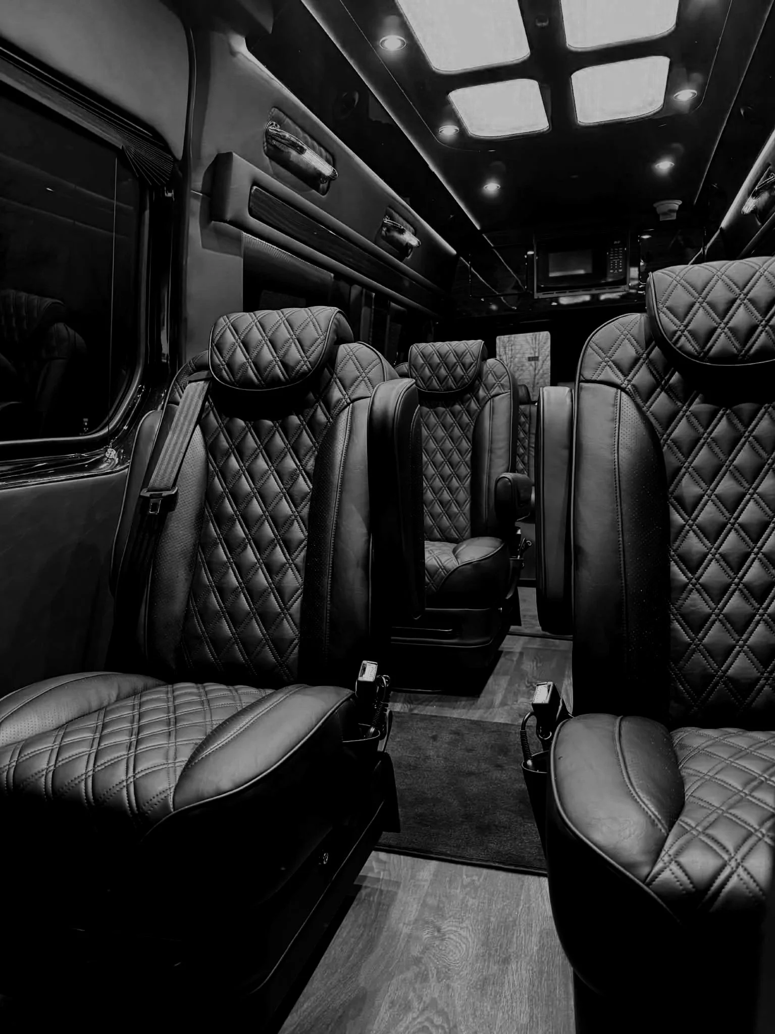 Black leather quilted seats inside a luxury bus or van, with wooden flooring and a TV monitor on the ceiling.