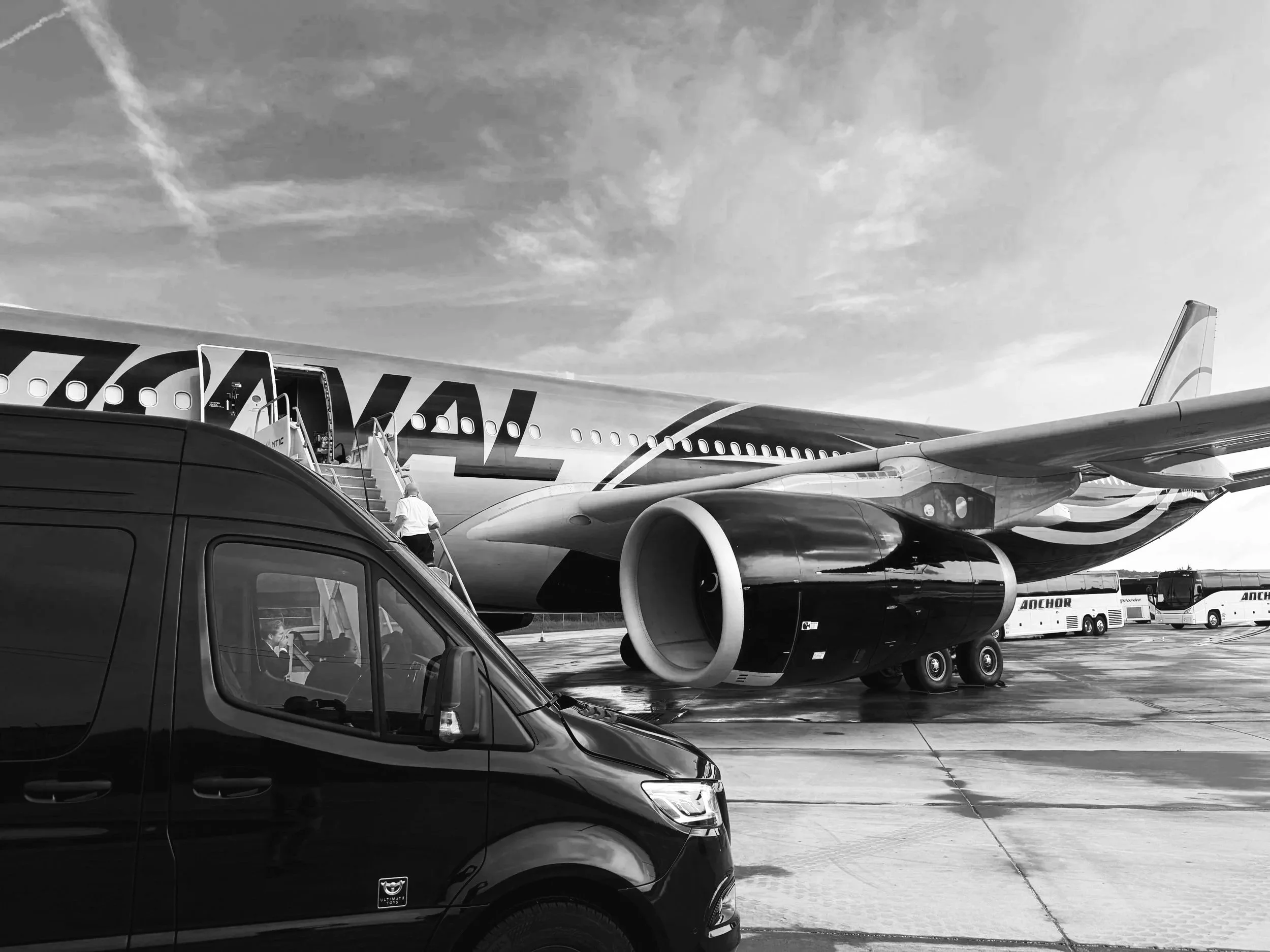 A large airplane is parked on the tarmac, with people boarding via stairs. In the foreground, a black van is visible, and in the background, there are buses and additional ground equipment. The photo is taken outdoors under a sky with scattered clouds.
