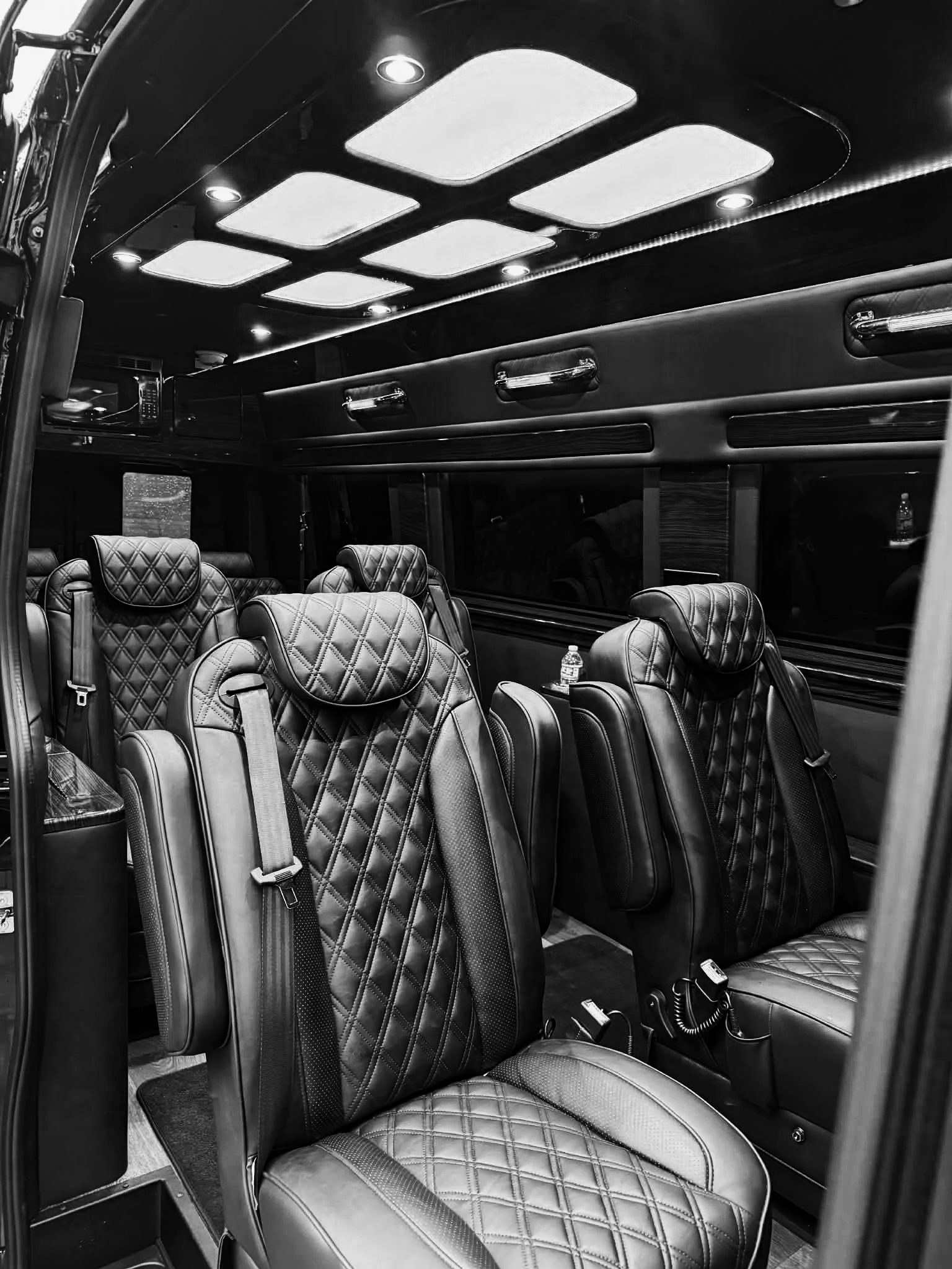 Black and white photo of luxury bus seating with quilted leather seats, headrests, and seatbelts, inside a bus at night.