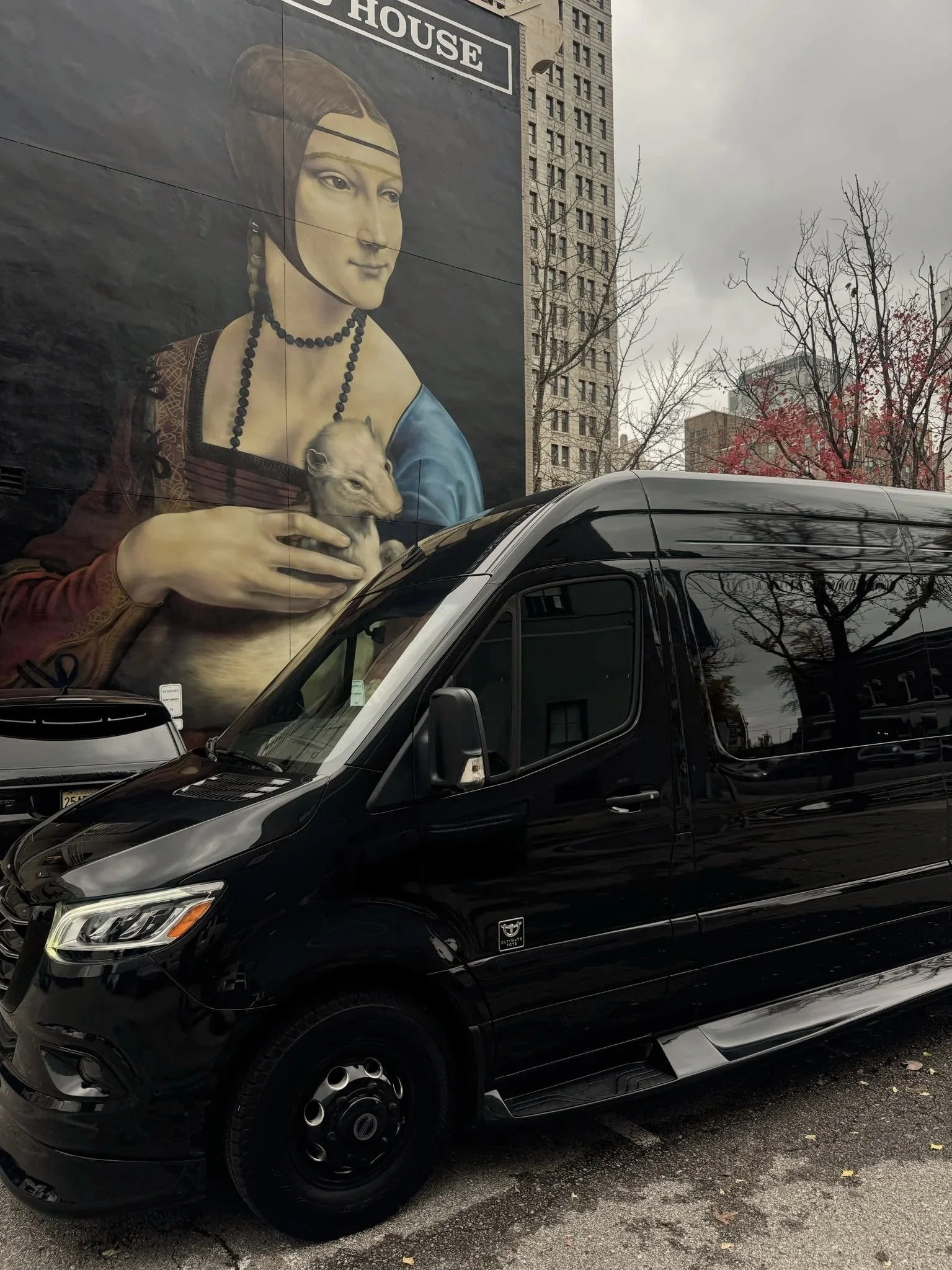 A large mural of a woman with historical clothing holding a small dog, painted on the side of a building, with a black van parked in front.