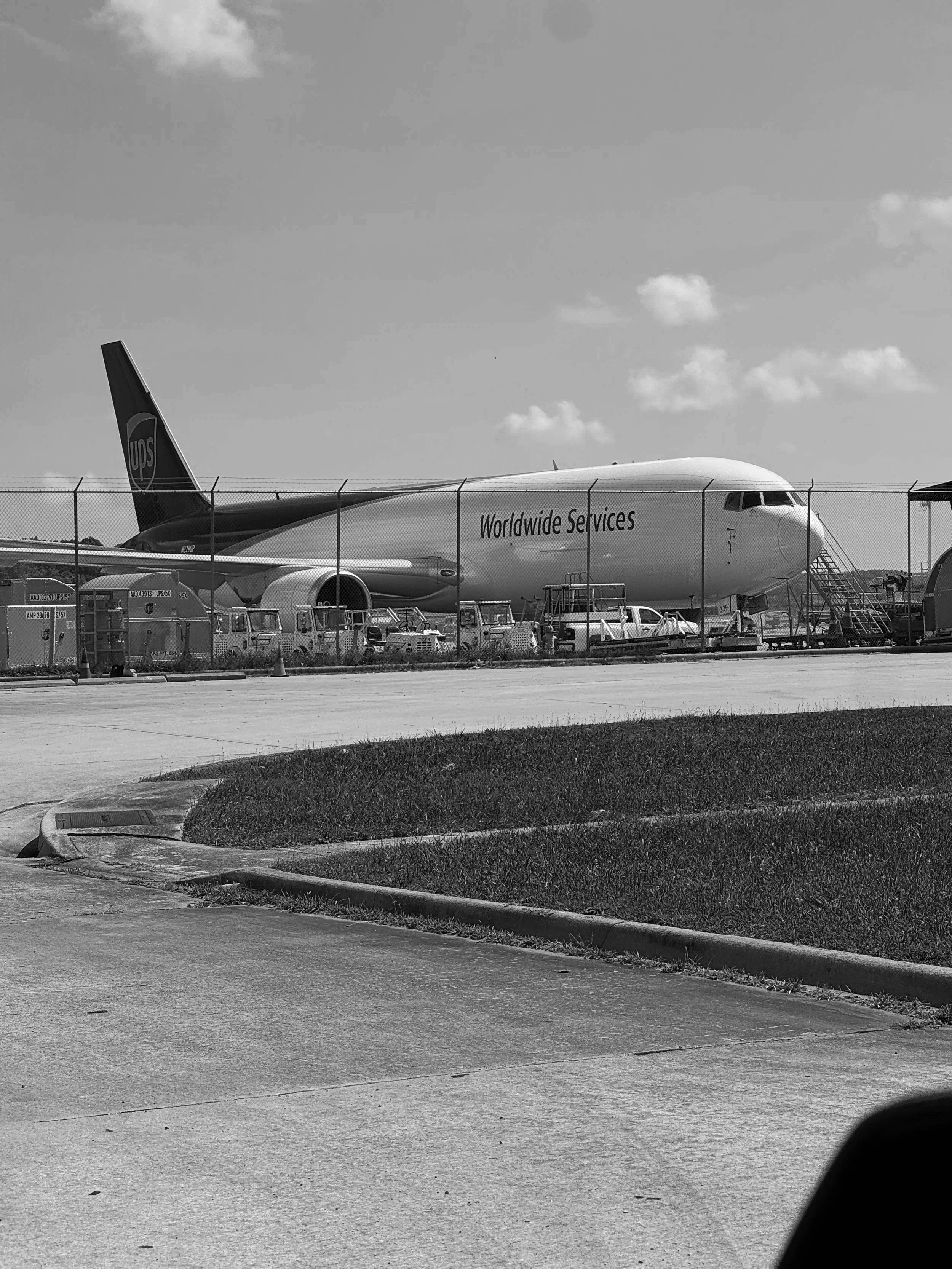 A large airplane on the tarmac behind a chain-link fence, with vehicles in front, under a partly cloudy sky.