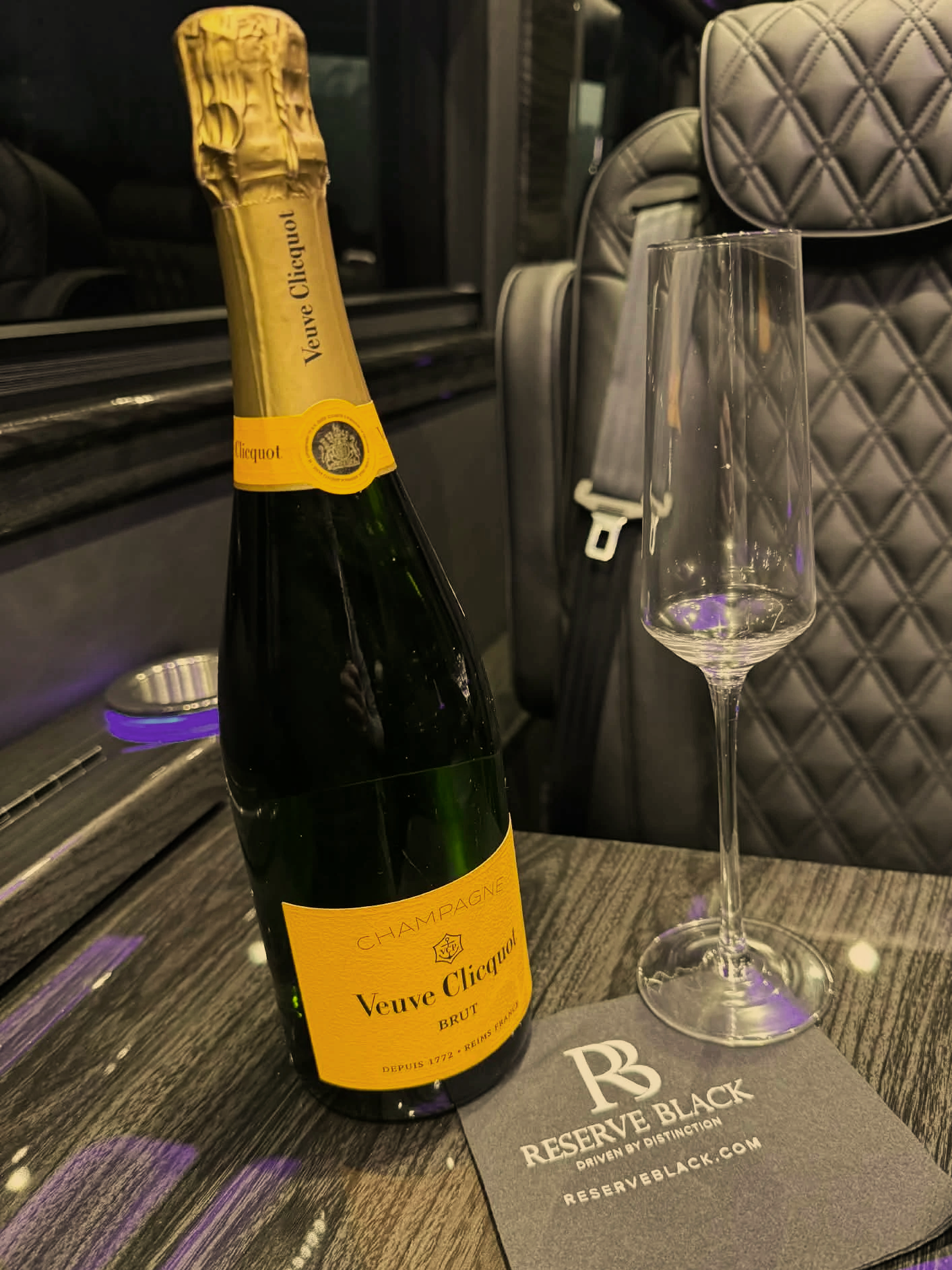 A bottle of Veuve Clicquot champagne and a champagne flute on a table with a Reserve Black coaster inside an airport lounge.